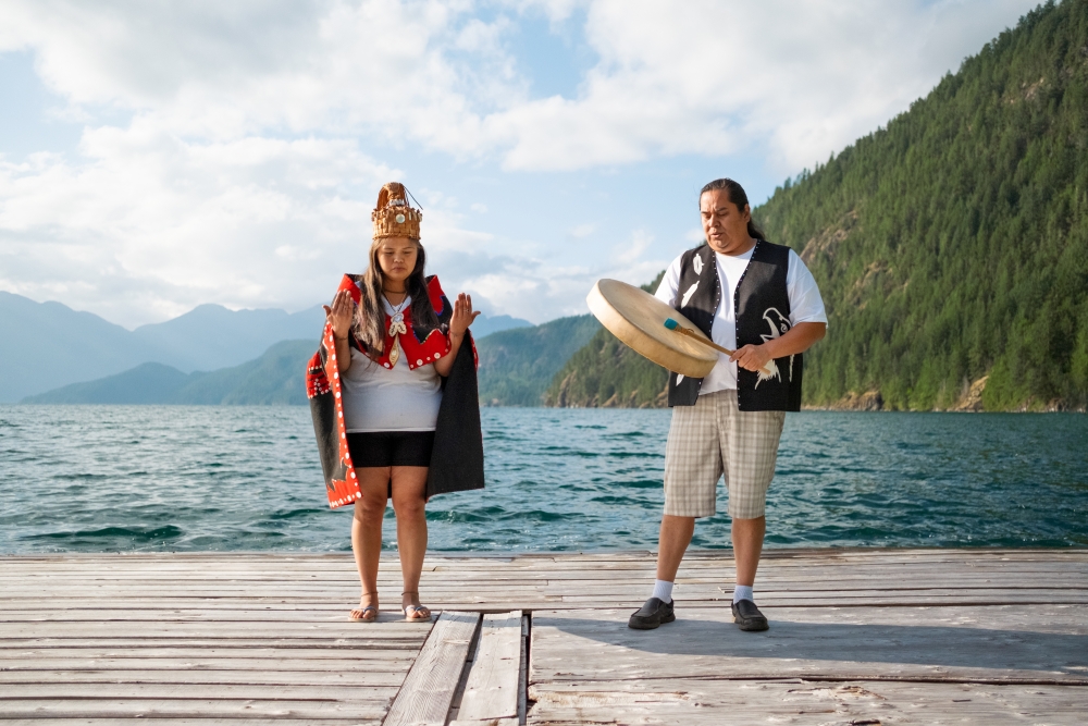 Two Indigenous people in British Columbia Canada at Desolation Sound stand on a dock by a lake and mountains, one is playing an instrument and the other has their hands raised